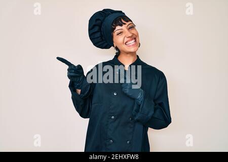 Schöne brunettte Frau trägt professionelle Koch Uniform lächelnd und Blick auf die Kamera zeigt mit zwei Händen und Fingern zur Seite. Stockfoto