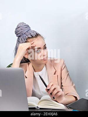 Büroarbeit, müde, überfordert. Porträt von müde junge moderne Frau mit Laptop im Büro trägt rosa Blazer und kanekolon Zöpfe. Kopfschmerzen, SED Stockfoto