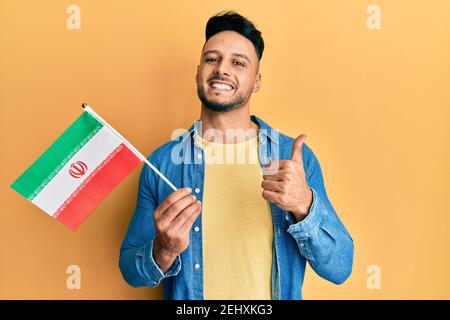 Junger arabischer Mann hält iran Flagge lächelnd glücklich und positiv, Daumen nach oben tun ausgezeichnet und Zustimmung Zeichen Stockfoto