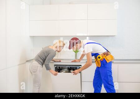 Junge Frau Schaut Auf Männlich Techniker Überprüfung Spülmaschine In Der Küche Stockfoto