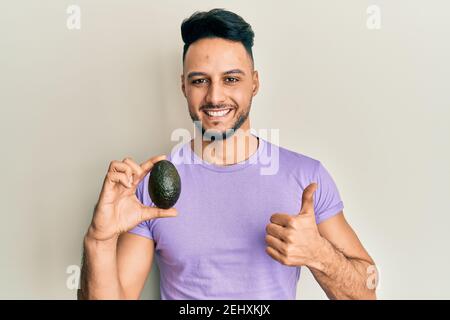 Junger arabischer Mann hält Avocado lächelnd glücklich und positiv, Daumen nach oben tun ausgezeichnet und Zustimmung Zeichen Stockfoto