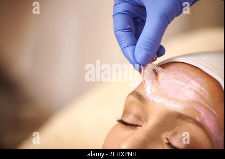 Entfernen der rosa Maske nach der Reinigung des Gesichts. Nahaufnahme. Der Ästhetiker entfernt die kosmetische Maske vom Gesicht eines entspannten Klienten Stockfoto