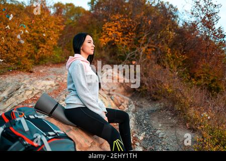 Eine junge kaukasische Frau sitzt auf einem Steinvorsprung und bewundert die Natur. Daneben ist ein Rucksack. Das Konzept von Wandern und Reisen. Stockfoto