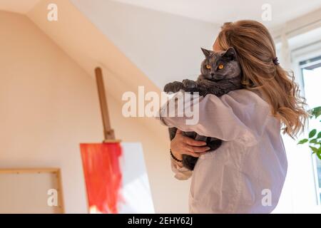 Frau hält ihre niedliche Katze im Kunststudio. Künstlerin versuchen, die Inspiration vor dem Malen zu finden. Britische Kurzhaar Katze und Haustierbesitzer Stockfoto