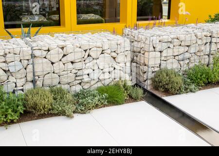 Garten mit einer mit Steinen gefüllten Gabion-Wand, gefrierendes Wasser im Garten, gepflasterte Terrasse - Pflanzen von mediterranen Kräutern an der Grenze nach Großbritannien Stockfoto