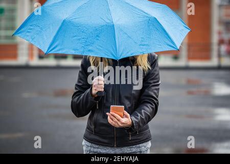 Frau mit Regenschirm hält Smartphone und steht im Regen auf der Straße. Wireless Internet Verbindung auf Handy in der Stadt Stockfoto