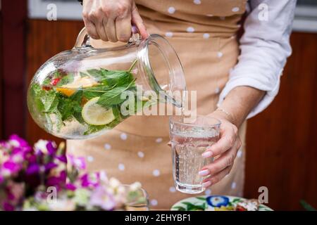 Frau gießt kaltes Mineralwasser mit Minzkräutern und Zitrone aus dem Krug in Trinkglas Stockfoto