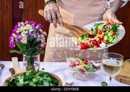 Frau mit Schürze Kochen frischen Gemüsesalat. Servieren von Lebensmitteln mit Holzlöffel auf Glas Schüssel auf dem Tisch. Stockfoto