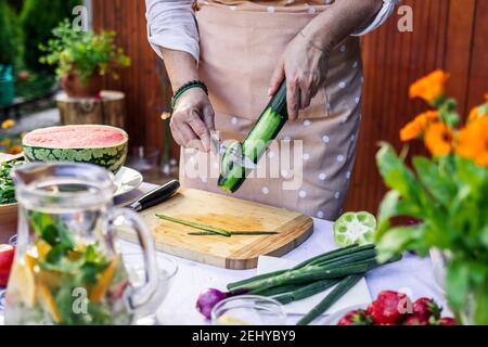 Frau schält Gurke auf dem Tisch. Frischen Gemüsesalat für Gartenparty. Vorbereitung von gesunden vegetarischen Speisen im Freien Stockfoto