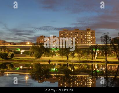 Stadtbild am Abend. Beleuchtete Häuser, Wege im Stadtpark, verschwommene Spur des Zuges, die sich im Stadtteich gegen den hellen Abendhimmel spiegelte Stockfoto