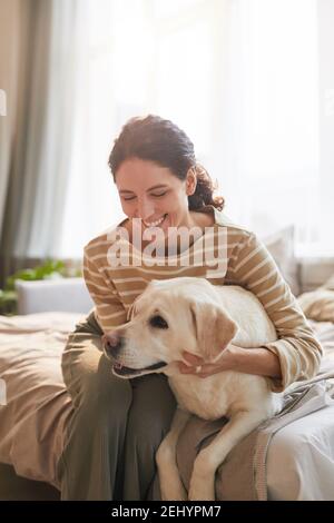 Vertikale warm getönten Porträt von lächelnden jungen Frau umarmenden Hund Liegen auf dem Bett in gemütlichen Innenraum von Sonnenlicht beleuchtet Stockfoto