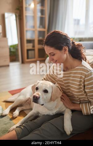 Vertikale warm getönten Porträt von lächelnden jungen Frau umarmenden Hund Während Sie auf dem Boden sitzen in gemütlicher Inneneinrichtung Stockfoto