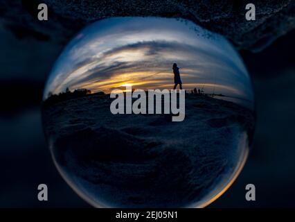 Surendorf, Deutschland. Februar 2021, 20th. In einer Glaskugel spiegeln sich Spaziergänger, die am Strand in Surendorf im Sonnenuntergang an der Eckernförder Bucht spazieren. (Kamera um 180 Grad gedreht) Quelle: Axel Heimken/dpa/Alamy Live News Stockfoto