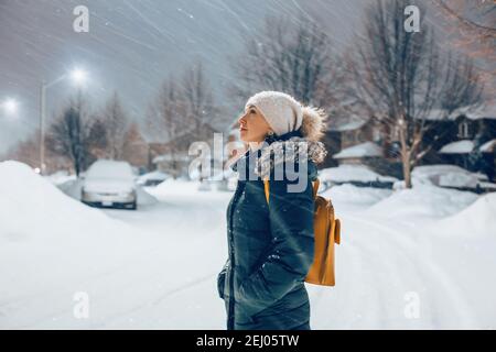 Frau in blauer Jacke Winterkleidung und Hut zu Fuß im Freien unter Schnee. Frau, die unter der Straßenbeleuchtung steht und den fallenden Schnee in der Nacht betrachtet Stockfoto