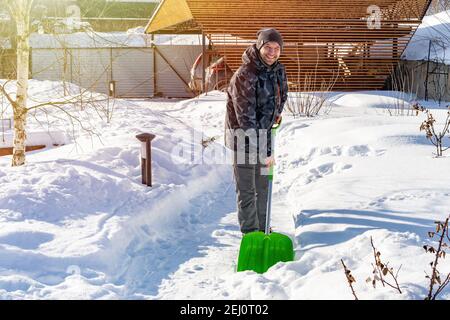 Ein Erwachsener lächelt Mann reinigt Schnee von den Spuren mit einer Schaufel nach starkem Schneefall an einem sonnigen Tag. Schneeräumung im Winter aus dem Garten. Hoher Schnee Stockfoto