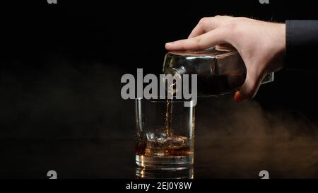 Der Barkeeper gießt goldenen Whiskey, Cognac, Brandy aus der Flasche in das Trinkglas mit Eiswürfeln auf schwarzem dunklen Hintergrund. Barkeeper Gießen leckeren Alkohol Cocktail Drink Bourbon, Rum in Zeitlupe. Nahaufnahme Stockfoto