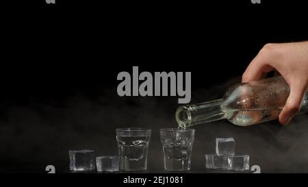 Barkeeper gießt vor schwarzem Hintergrund zwei Schüsse Wodka mit Eiswürfeln aus einer Flasche in Gläser. Barkeeper gießen klar transparent Alkohol trinken Wodka Tequila in Shot-Glas Stockfoto