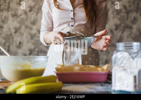 Die weiblichen Hände sieben das Mehl aus dem Sieb nach dem Teig in die Form für das Backen. Alte Küchengeschirr mit Mehl, Milch und Eiern im Vordergrund. Authentischer rustikaler Stil Stockfoto