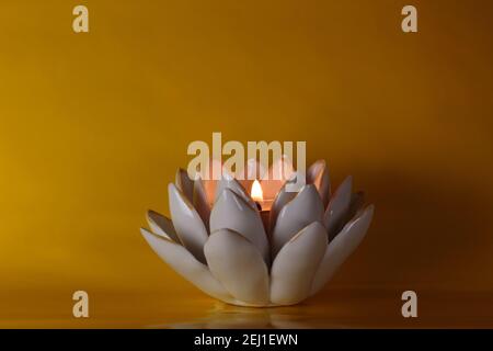 Brennende Teelicht mit Flamme in einem keramischen Lotusblumen Kerzenhalter auf einem tiefgelben Hintergrund mit dramatischer, romantischer Beleuchtung und Kopierraum Stockfoto