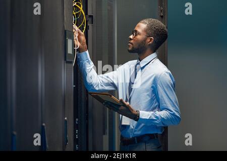 Grafik Seitenansicht Porträt eines afroamerikanischen Netzwerktechnikers, der Kabel im Serverschrank während der Arbeit mit Supercomputern im Rechenzentrum verbindet, c Stockfoto