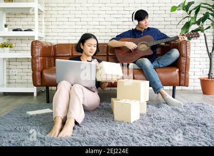 Junge Liebhaber verbringen Zeit zusammen auf Urlaub im Wohnzimmer. Asiatischer Mann mit Kopfhörern spielt eine akustische Gitarre, während das Mädchen das Paket überprüfen Stockfoto