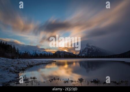 Langzeitbelichtung Sonnenaufgang Landschaft von Bergen mit Schnee reflektiert auf dem Wasser während eines kalten morgendlichen Winters unter einem bewölkten Himmel, Mount Rundle, Kanada Stockfoto