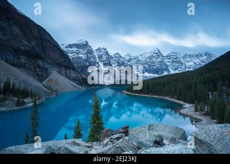 Sonnenaufgang in den Bergen mit Schnee während eines bewölkten Herbstmorgens, blauer ruhiger See mit Reflexen, grünen Bäumen und grauen Felsen, Kanada Stockfoto