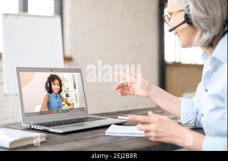 Eine ältere, grauhaarige Geschäftsfrau im Headset, die virtuelle Meetings mit dem Kunden führt, Online-Geschäftsvideoanruf über einen Laptop vom Heimbüro aus. Senior Stockfoto