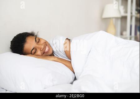 Multiethnische Frau schlafen in reinem Bett zu Hause, liegen auf weichen frischen Kissen, lächelnd, Seitenansicht, beobachten angenehmen Traum, tragen in weiß einfach Stockfoto