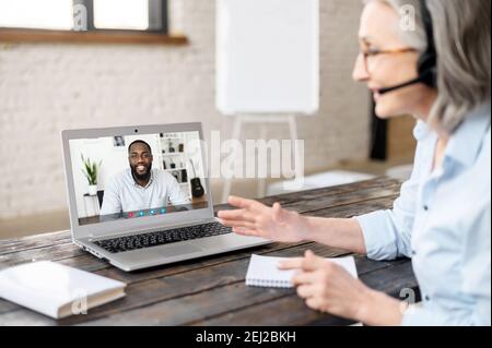 Ältere grauhaarige Therapeutin im Headset Blick auf den Bildschirm, im Gespräch mit einem jungen schwarzen Mann, mit virtuellen Beratung mit Client, Online-Video Stockfoto