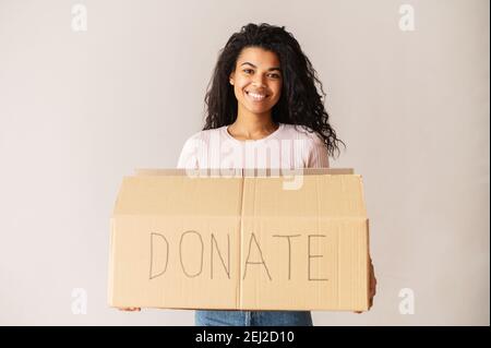 Freundlich lächelnde afroamerikanische Freiwillige mit lockigen dunklen Haaren hält einen Karton und spendet ihre Sachen für wohltätige Zwecke, für Waisenkinder und Obdachlose Stockfoto
