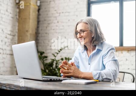 Ältere Geschäftsfrau sitzt am Schreibtisch, Blick auf Webcam und im Gespräch mit Kunden online oder beobachten Tutorial, ältere weibliche Büroangestellte oder Stockfoto