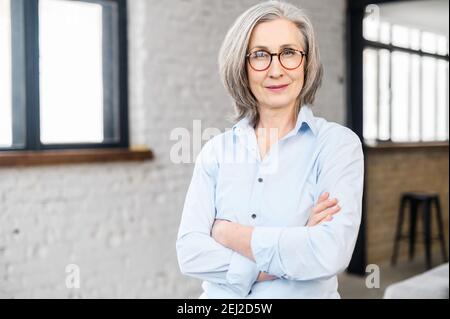 Porträt einer älteren reifen grauhaarigen professionellen Frau, Führer, Coach, ältere Geschäftsfrau in Brille stehend mit gekreuzten Armen gefaltet in Stockfoto
