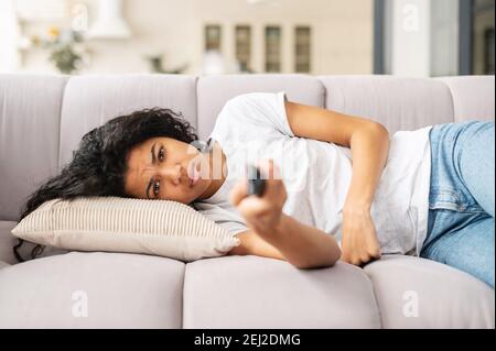 Aufgeregt junge Birazialfrau liegt auf einem weißen Sofa mit einer Fernbedienung in der Hand, kann nichts Interessantes im fernsehen finden, auf einer Selbstisolation, zu Hause Stockfoto