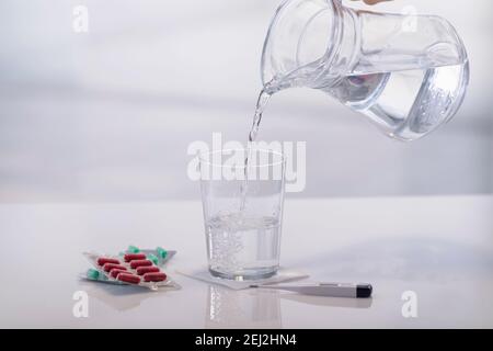 Nahaufnahme einer Hand, die Wasser in ein Glas gießt, das von Medikamentenkapseln, Blasen und einem Digitalthermometer umgeben ist. Gesundheitskonzept. Stockfoto