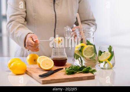 Weibliche Hand hält einen tropfenden Honigtaucher über einem Honigglas auf einem Holzschneidetisch mit Zitronen. Selektiver Fokus. Hausgemachte Heilmittel Konzept. Stockfoto
