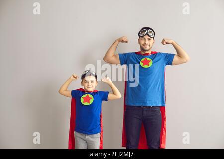 Kraftvoller Vater mit Sohn im Superhelden-Kostüm, der Muskeln vor der Kamera auf grauem Hintergrund zeigt. Stockfoto