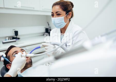 Zahnarzt trägt Gesichtsmaske lächelnd, während eine zahnärztliche Behandlung auf männlichen Patienten. Zahnärzte arbeiten an den Zähnen eines Patienten in der Klinik. Stockfoto