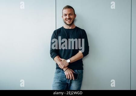 Porträt eines selbstbewussten Geschäftsmannes in Casuals, der in die Kamera blickt und lächelt. Der Geschäftsmann steht im Büro an der Wand. Stockfoto