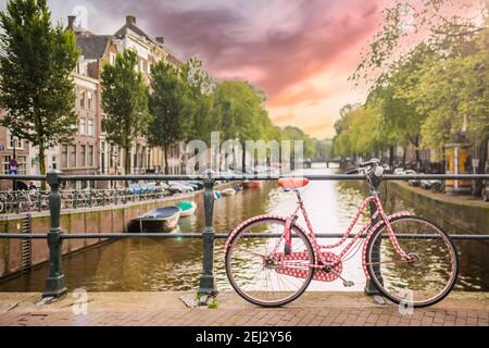 (Selektiver Fokus) atemberaubende Aussicht auf ein pinkes Fahrrad, das auf einer Brücke über einen der vielen Kanäle Amsterdams geparkt ist. Schöner Sonnenuntergang im Hintergrund. Stockfoto