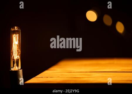 (Selektiver Fokus) Vintage-Glühbirne auf leerem Holztisch, beleuchtet von einem warmen Licht, schwarzem Hintergrund mit einer verschwommenen Lichtkette. Stockfoto