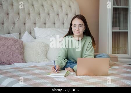 Nettes Teenager-Mädchen macht Hausaufgaben.Junges Mädchen trägt eine Brille schriftlich im Notebook, Online-Studium mit Laptop, Fernstudium, Selbstbildung Stockfoto