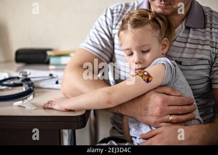 Impfung eines kleinen Mädchens in den Armen ihres Vaters in der Arztpraxis vor dem Coronavirus. Kinder lustige Klebstoff Gips. Impfstoff gegen Covid-1 Stockfoto