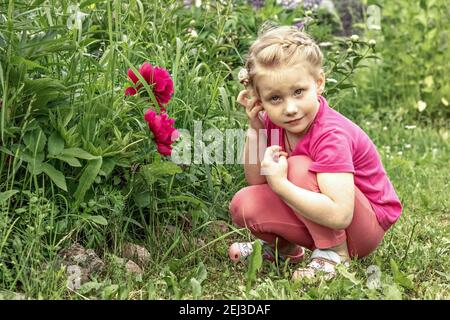 Das kleine Mädchen ist in Gedanken verloren, sitzt am Blumenbeet im Garten der rosa Pfingstrosen. Stockfoto