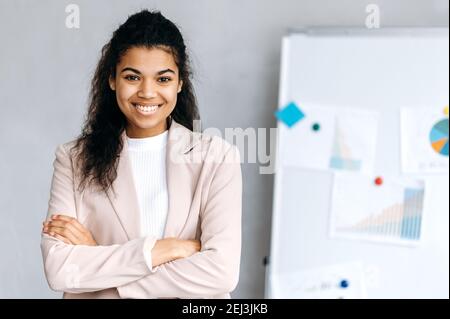 Porträt einer wunderschönen afroamerikanischen jungen Geschäftsfrau in stilvollem formellen Anzug, stehend mit gekreuzten Armen im Büro, schauen und lächeln freundlich auf die Kamera Stockfoto