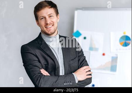 Porträt eines attraktiven kaukasischen jungen Geschäftsmannes, gekleidet in stilvollen formellen Anzug, stehend mit gekreuzten Armen im Büro, schauen und lächeln freundlich auf die Kamera Stockfoto