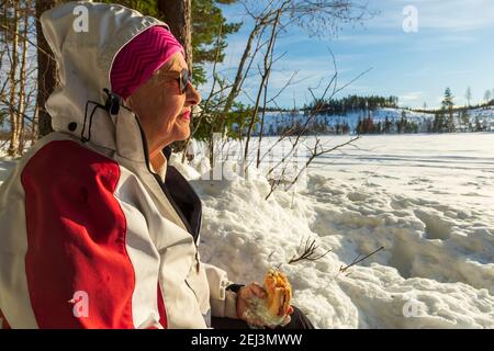Ältere Frau, die sich ausruhte, sitzt am Ufer eines gefrorenen Sees und hält ein Sandwich in der Hand, Bäume und einen blauen Himmel im Hintergrund, Bild von Vastre Stockfoto