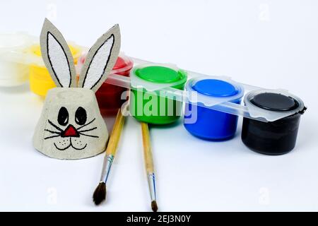 Osterhase aus Pappeibecher neben Pinsel und bunten Acryl-Bastelfarben. Weißer Hintergrund Stockfoto