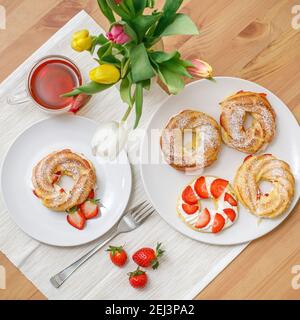 Profiteroles oder Cremepuffkuchen mit Schlagsahne und Erdbeeren, serviert auf dem Tisch mit Tulpenblüten Stockfoto