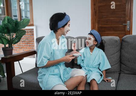 Eine Mutter und ihr kleines Mädchen lächeln in einer Creme Gesichtsmaske trägt ein Handtuch und Stirnband sitzen auf einem Stuhl Stockfoto